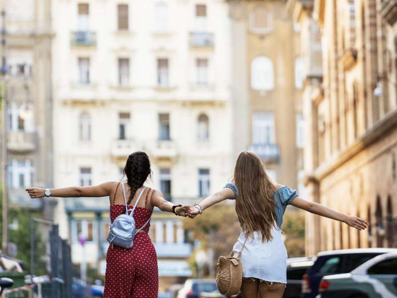 Back view of anonymous female friends in summer clothing holding hands and walking along street in old tourist town